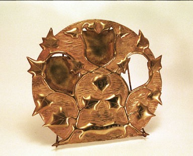 Round brass sheet cut-out with three openings among a garland of ivy. Souvenir&nbsp; and 1919 engraved on one of the garland loop.